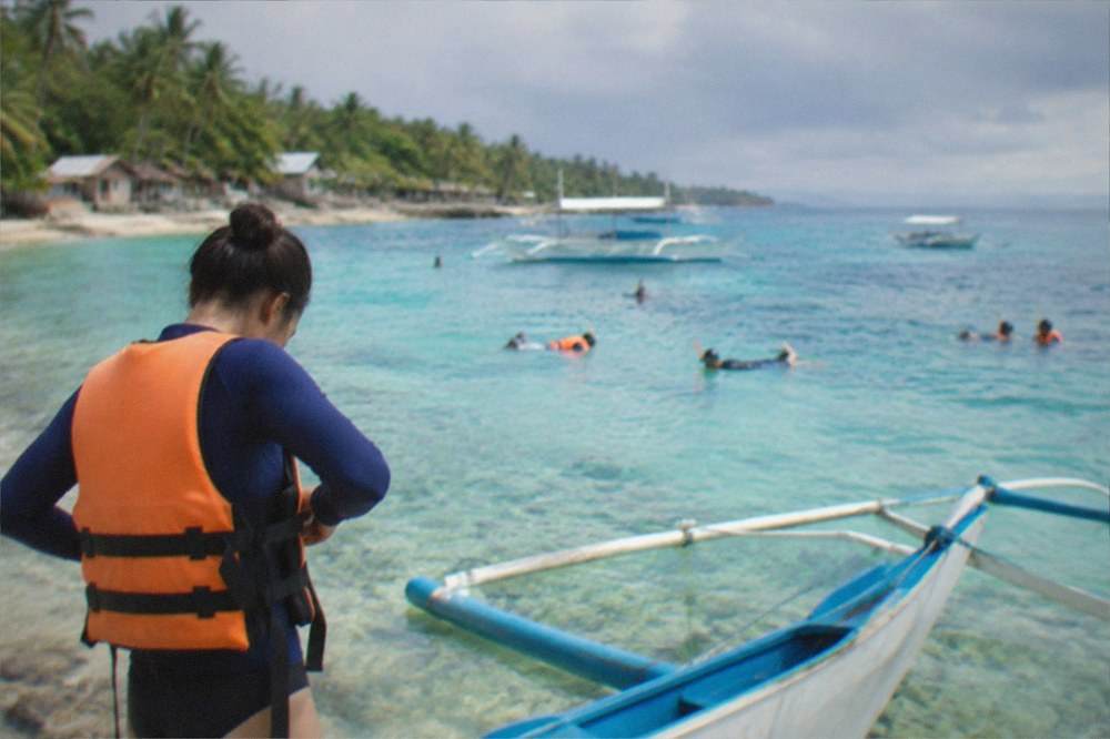 A visitor in a rash guard putting on a life jacket beside an outrigger boat at Oslob