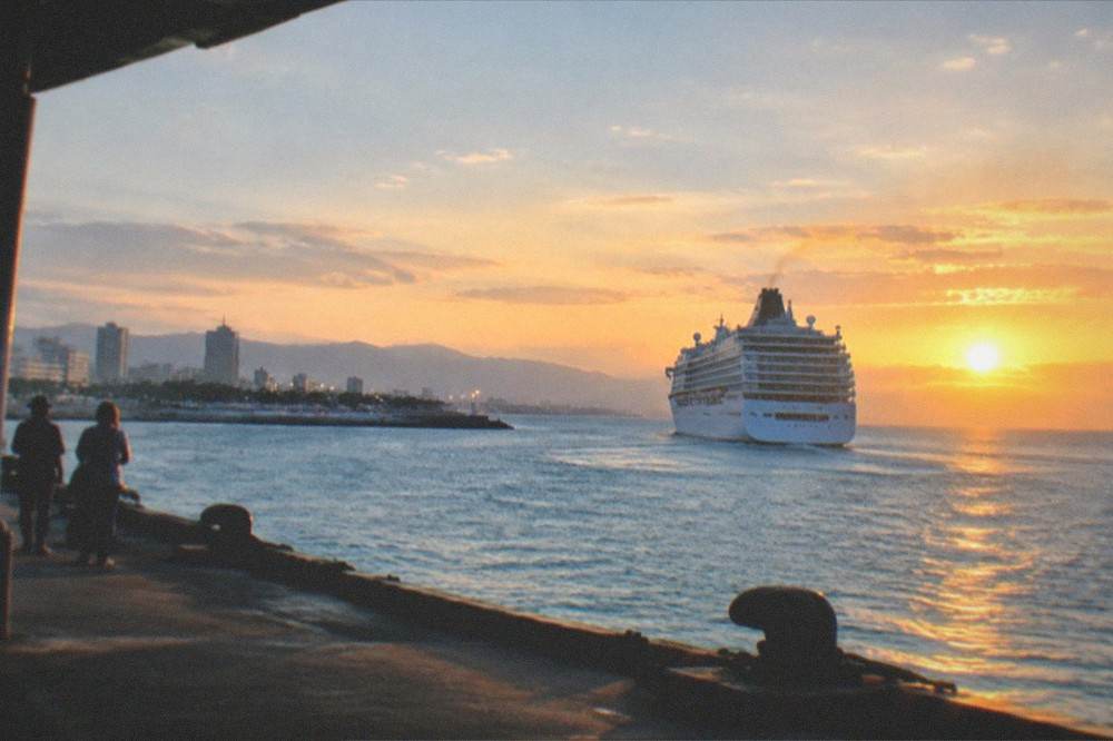A cruise ship departing a tropical island harbor at sunset with lights along the shore