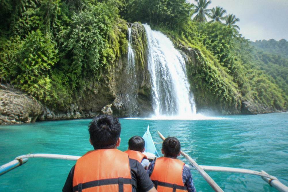 A family in life jackets on a small outrigger boat approaching a waterfall in southern Cebu