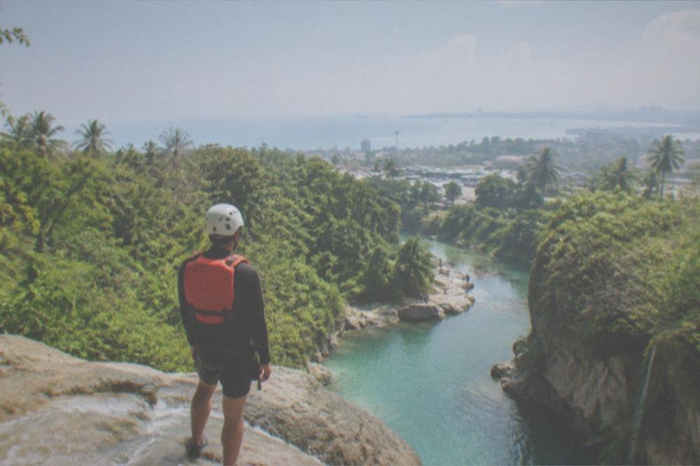 A hiker in a helmet and life jacket standing at the top of a canyon slide above a green river